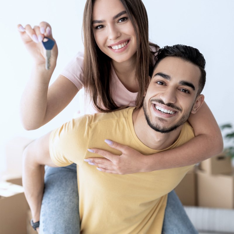 Cheerful Caucasian lady getting piggyback ride from her Arab boyfriend, showing house key indoors Cheerful Caucasian lady getting piggyback ride from her Arab boyfriend, showing house key indoors. Lovely multinational couple posing in new apartment on moving day